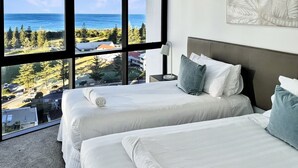Ein Hotelzimmer mit zwei Betten, einem großen Fenster mit Blick auf das Meer und Bäume, sowie einem modernen Kopfteil.