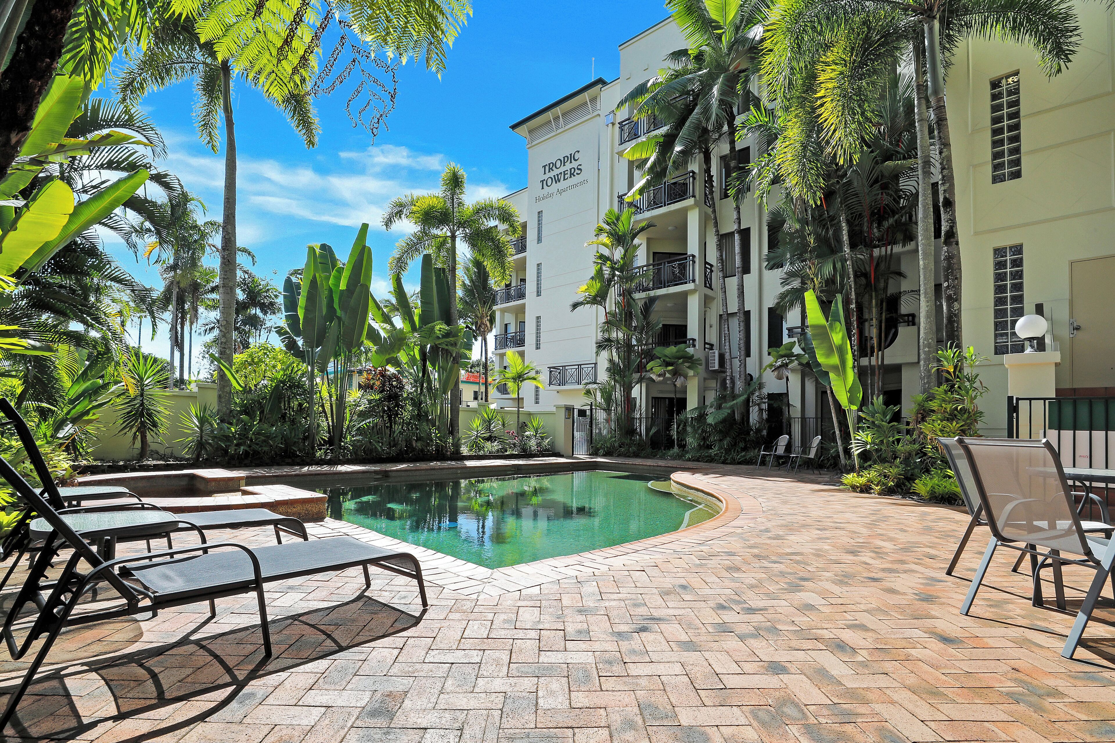 A pool area with lounge chairs, surrounded by tropical plants and a building labeled "TROPIC TOWERS."