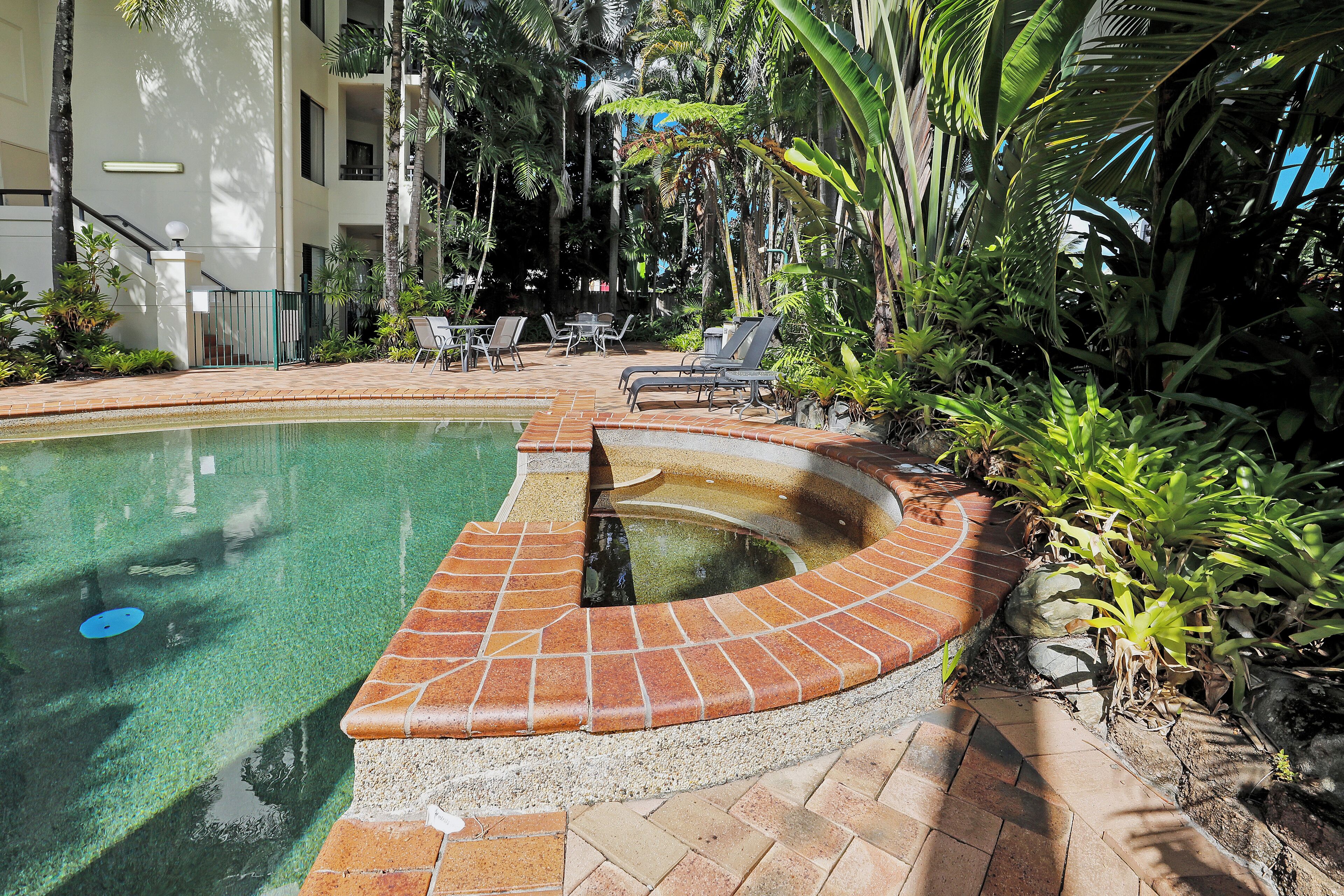 A pool with a brick edge, surrounded by lounge chairs and greenery.