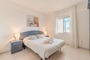 Uma cama bem arrumada com lençóis brancos e duas almofadas, uma mesa de cabeceira com abajur, e um quadro na parede.