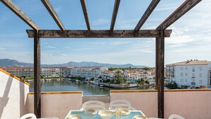 A balcony with a table set for dining, overlooking a river and a town with buildings and mountains in the distance. - Flateli Empuriabrava (Empuriabrava)