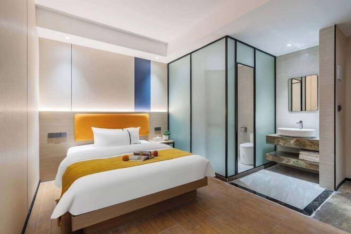 Habitación de hotel moderna con una cama grande, un separador de vidrio, un baño con lavabo y espejo, y piso de madera.
