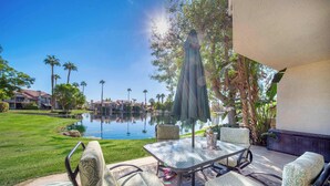 A patio with a glass table and wicker chairs, overlooking a pond and a well-maintained lawn.