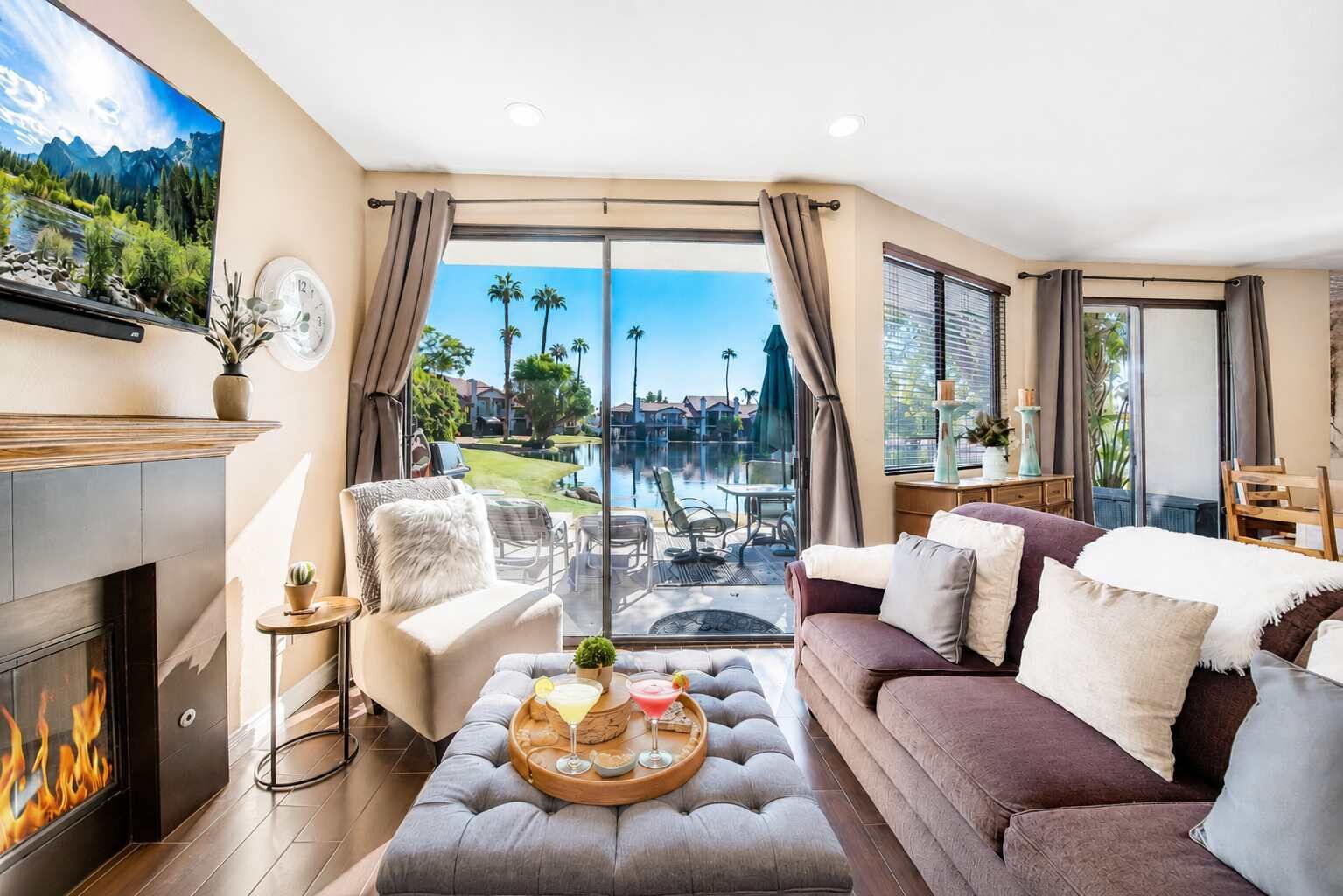 A living room with a fireplace, a sofa, a coffee table with drinks, and a view of a pool and mountains.