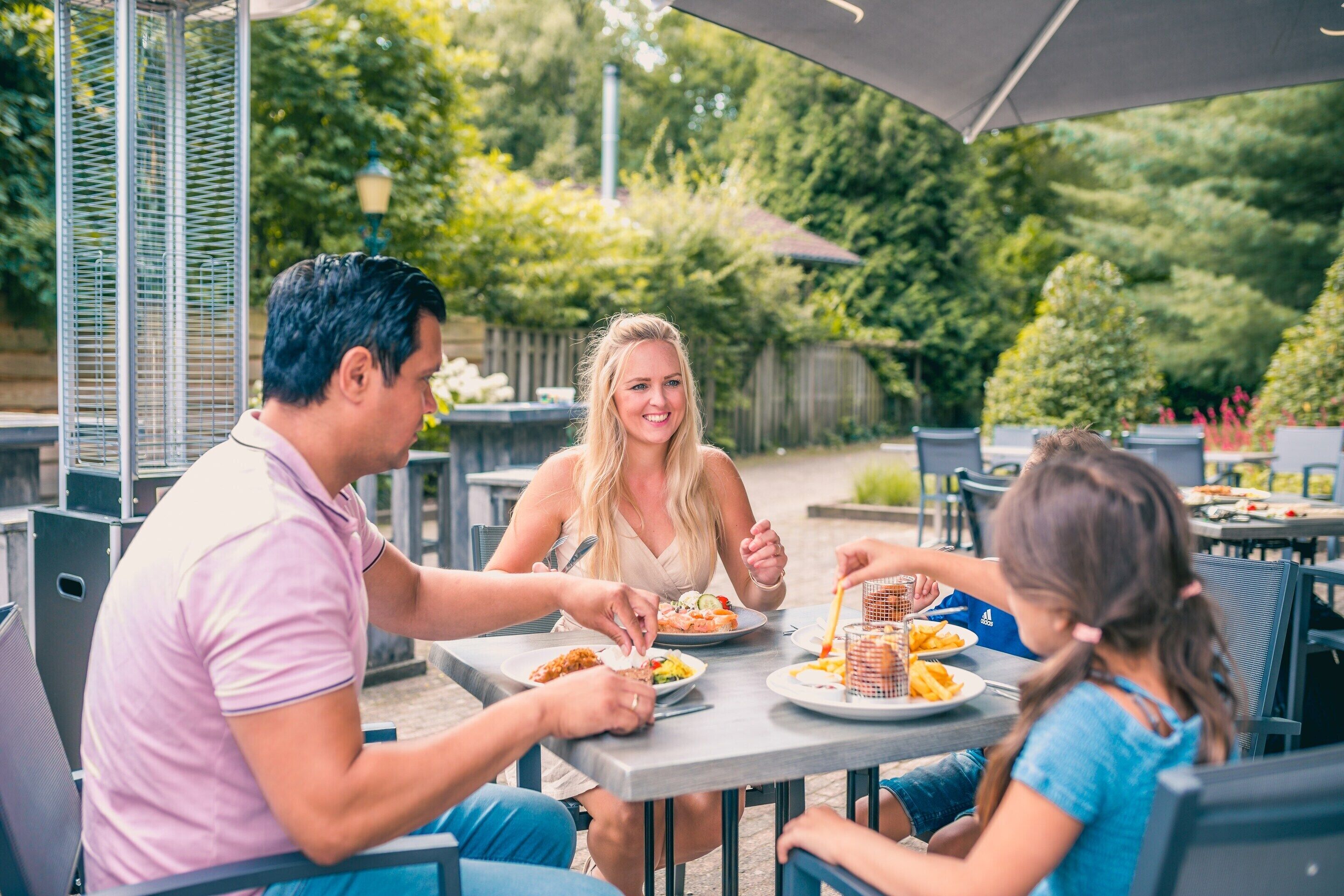 A family dining outdoors at a table with a metal grill, surrounded by greenery and a wooden fence.