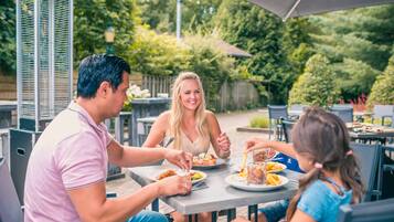 A family dining outdoors at a table with a metal grill, surrounded by greenery and a wooden fence.