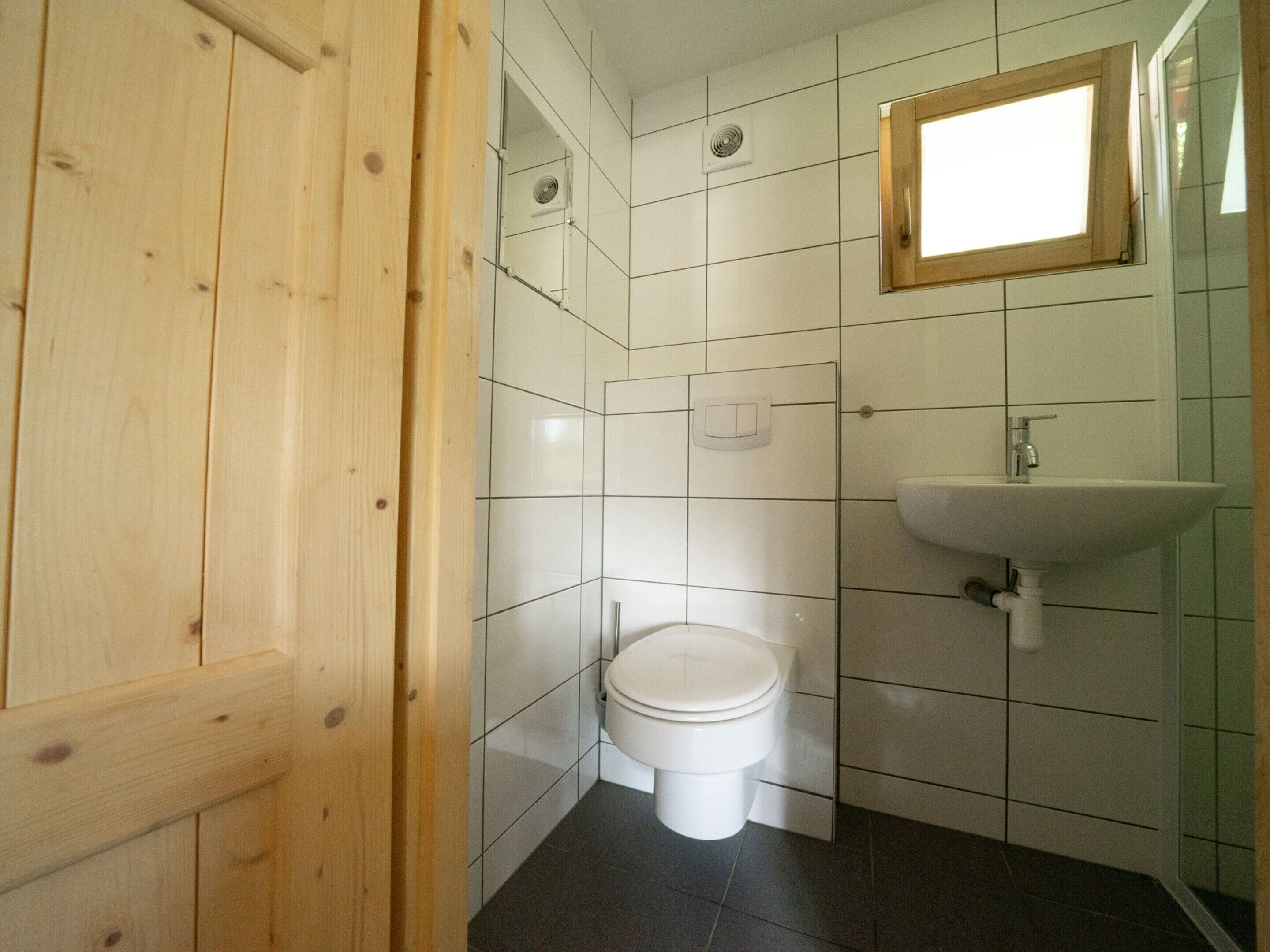 Un baño pequeño y limpio con azulejos blancos, una puerta de madera, un inodoro blanco, un lavabo y una ventana.