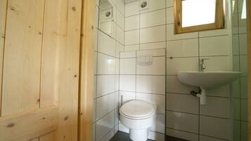 Un baño pequeño y limpio con azulejos blancos, una puerta de madera, un inodoro blanco, un lavabo y una ventana.