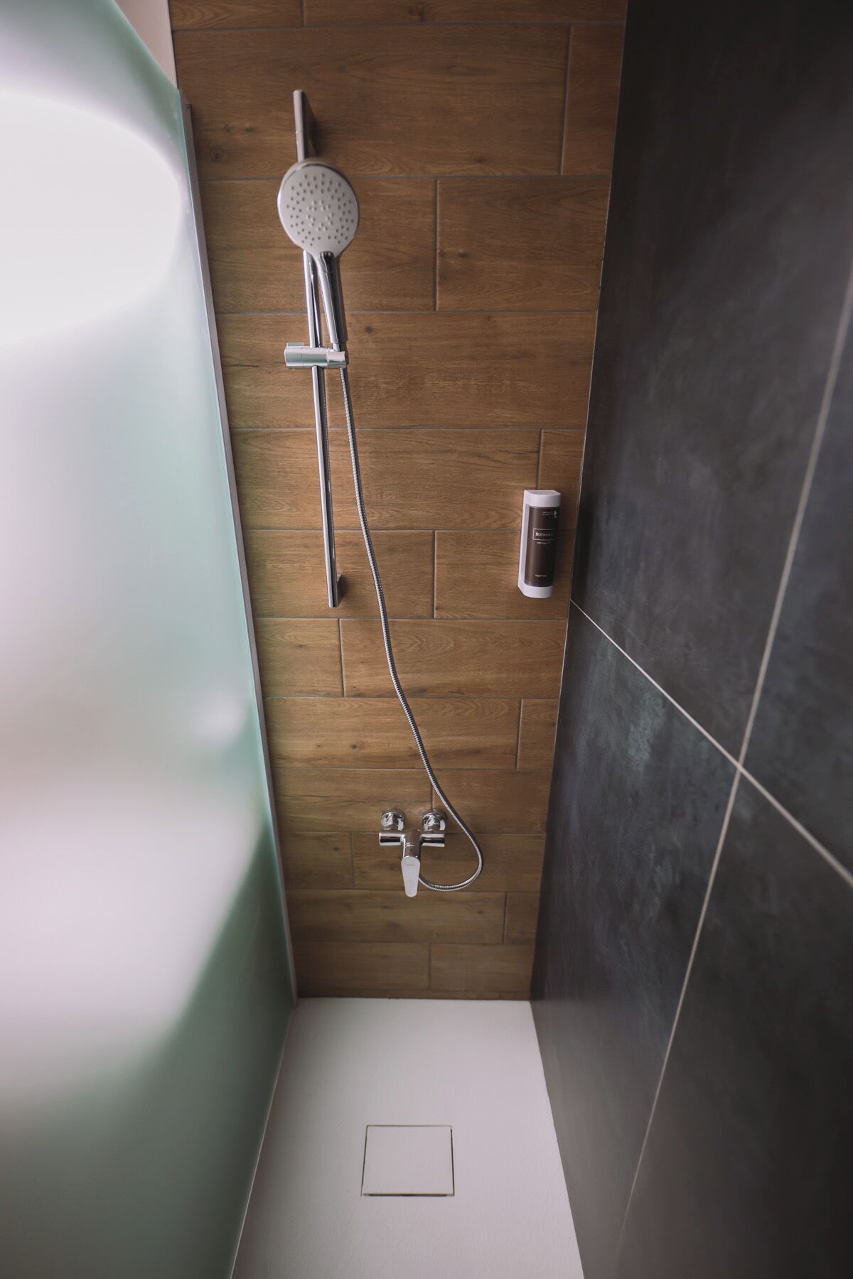 A shower with a handheld showerhead and a wall-mounted faucet, a soap dispenser, and a toilet in a bathroom with wooden and dark tiled walls.
