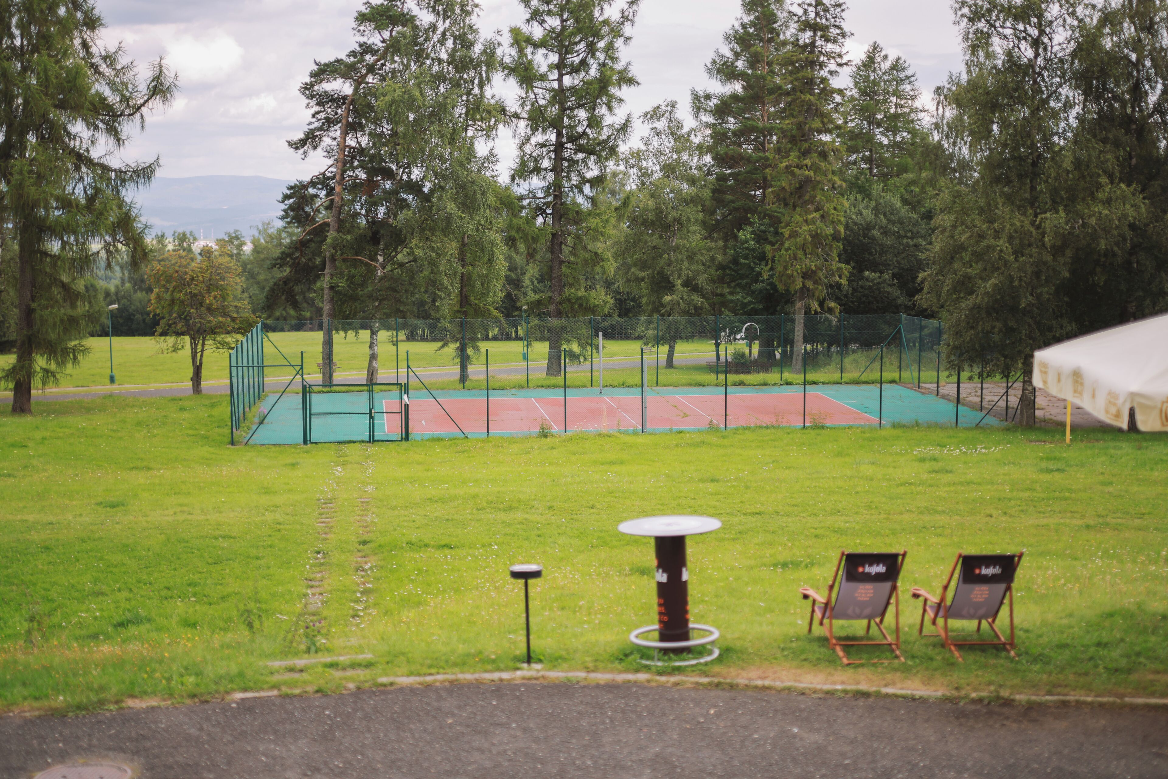 Una cancha de tenis con red, rodeada de árboles y un área verde. Hay dos sillas y una mesa en primer plano.