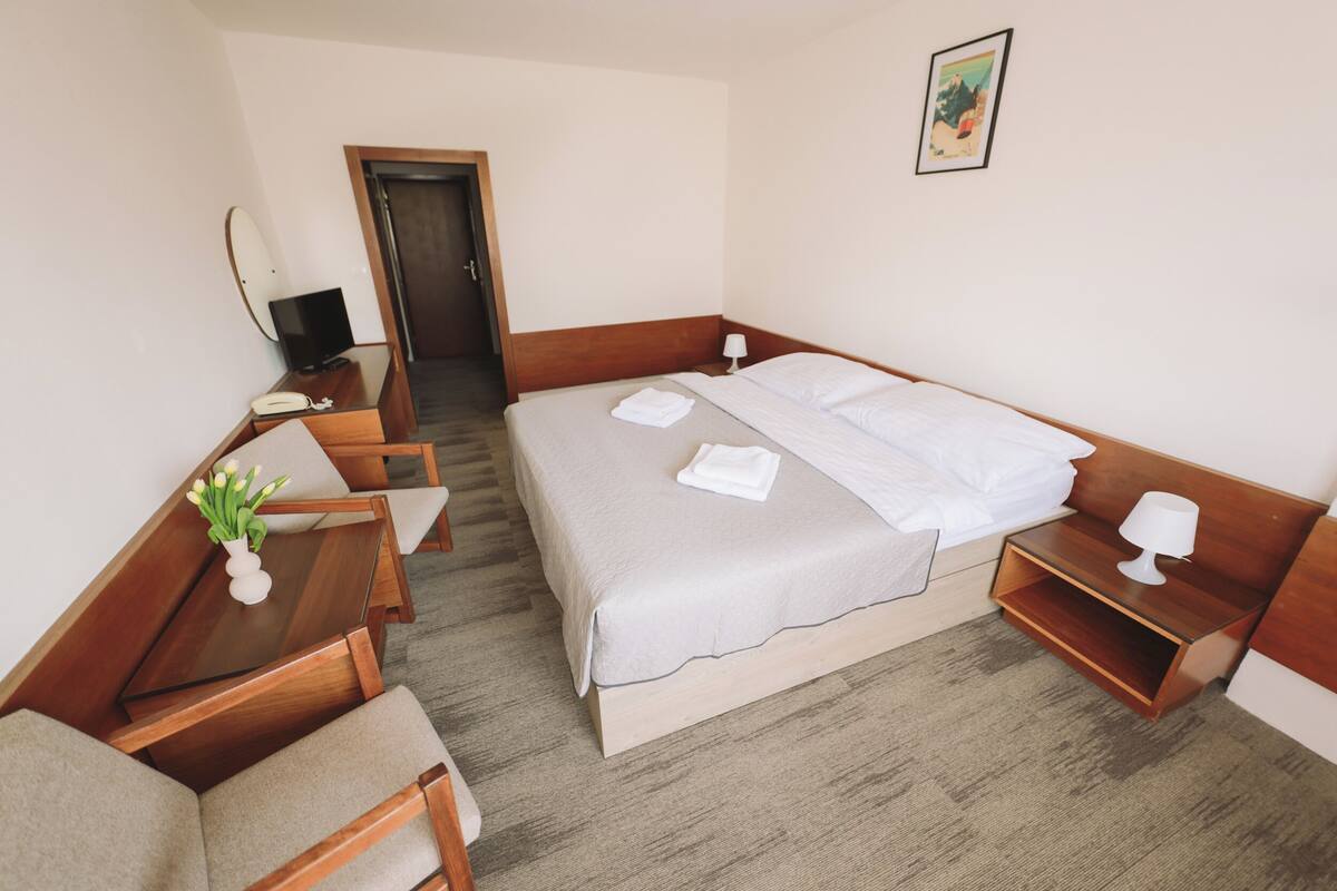 Quarto de hotel com cama, escrivaninha, poltrona, televisão e uma mesinha com um vaso de flores.