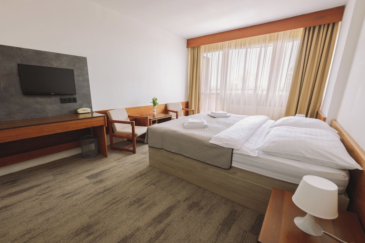 Quarto de hotel com cama, escrivaninha, cadeira, televisão e abajur.
