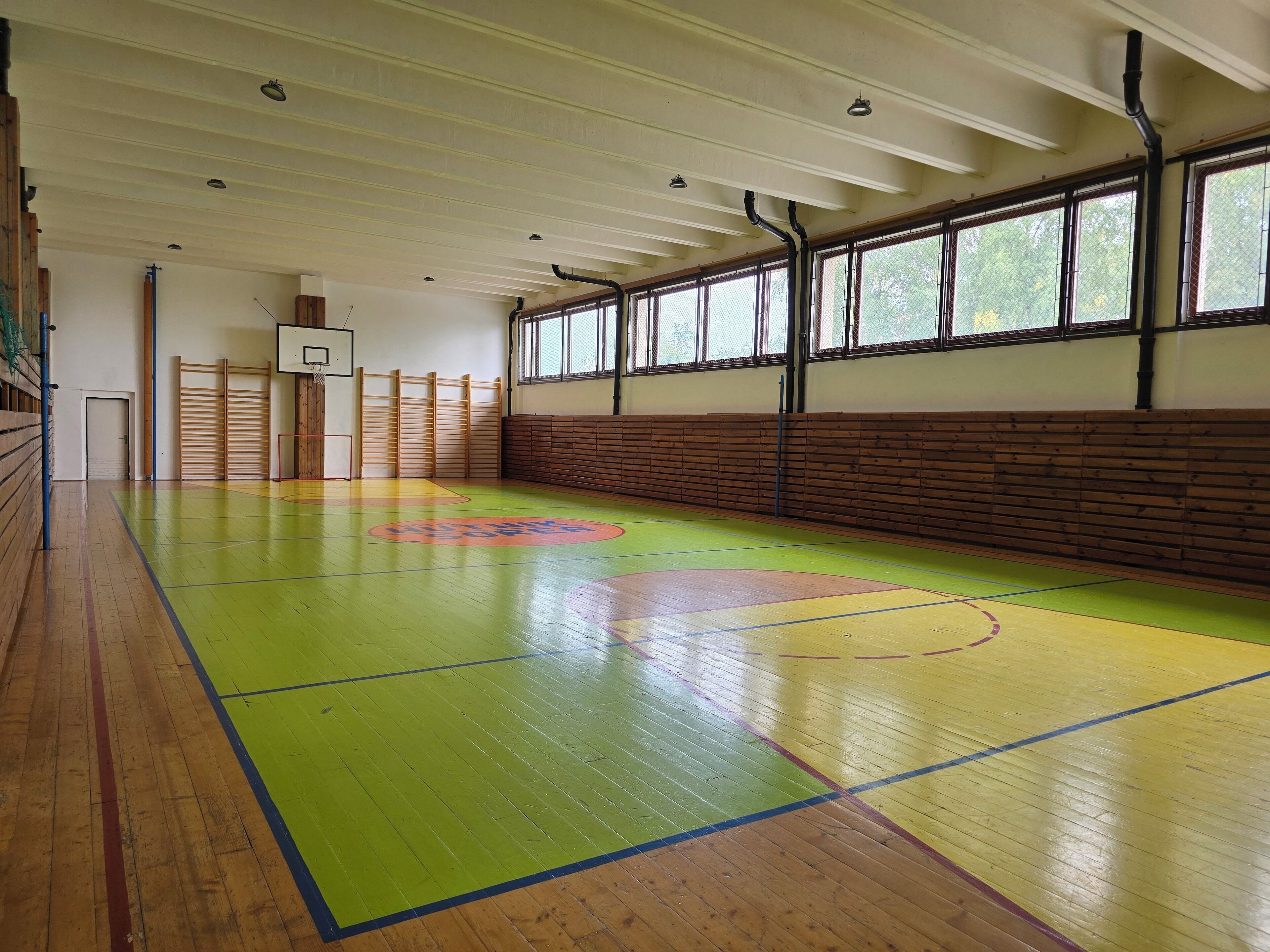 Una cancha de baloncesto interior con piso de madera, aro de baloncesto y pared de madera con ventanas.