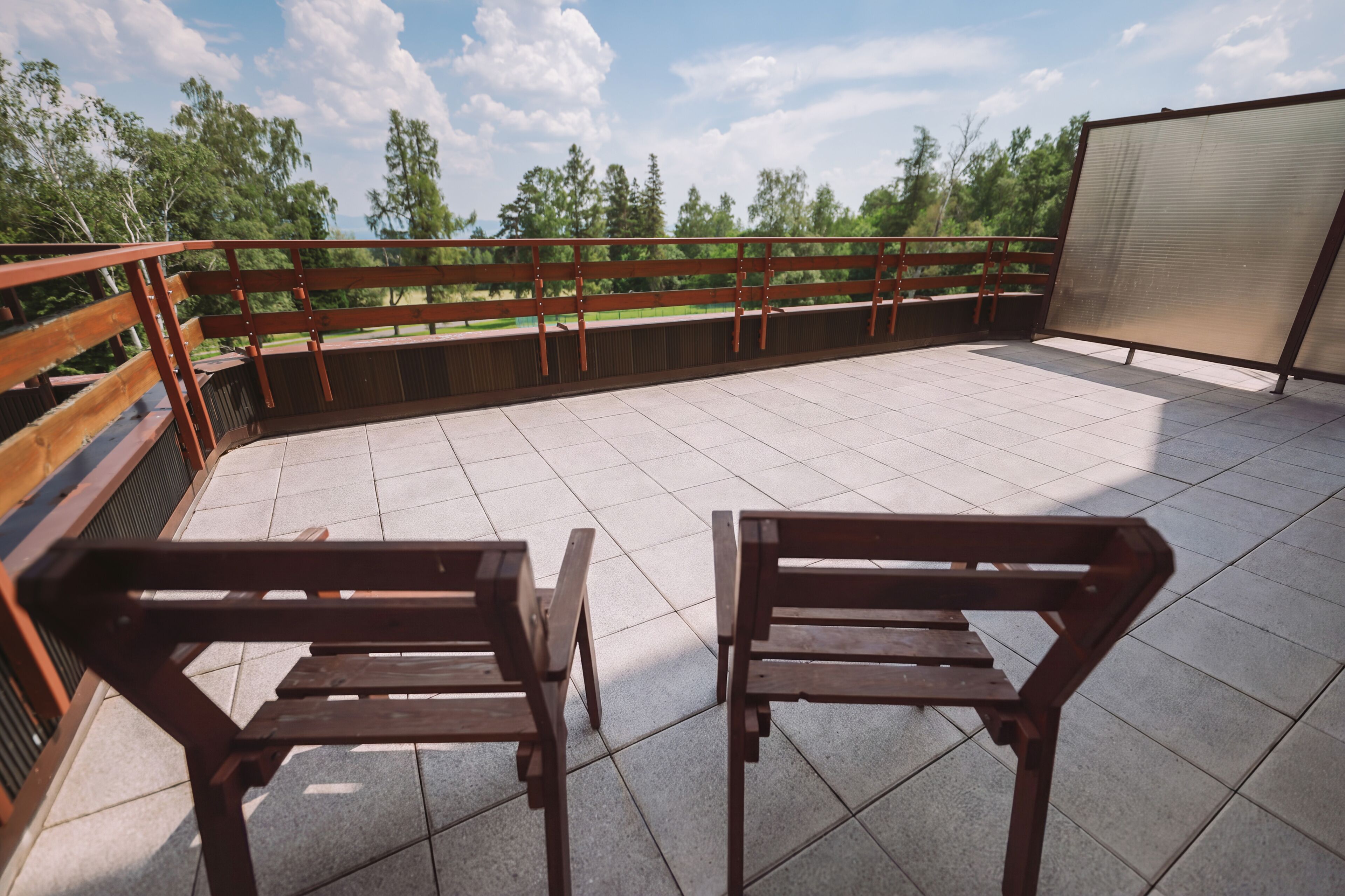 Eine Dachterrasse mit zwei Holzstühlen, einem Metallgeländer und Blick auf Bäume und Himmel.