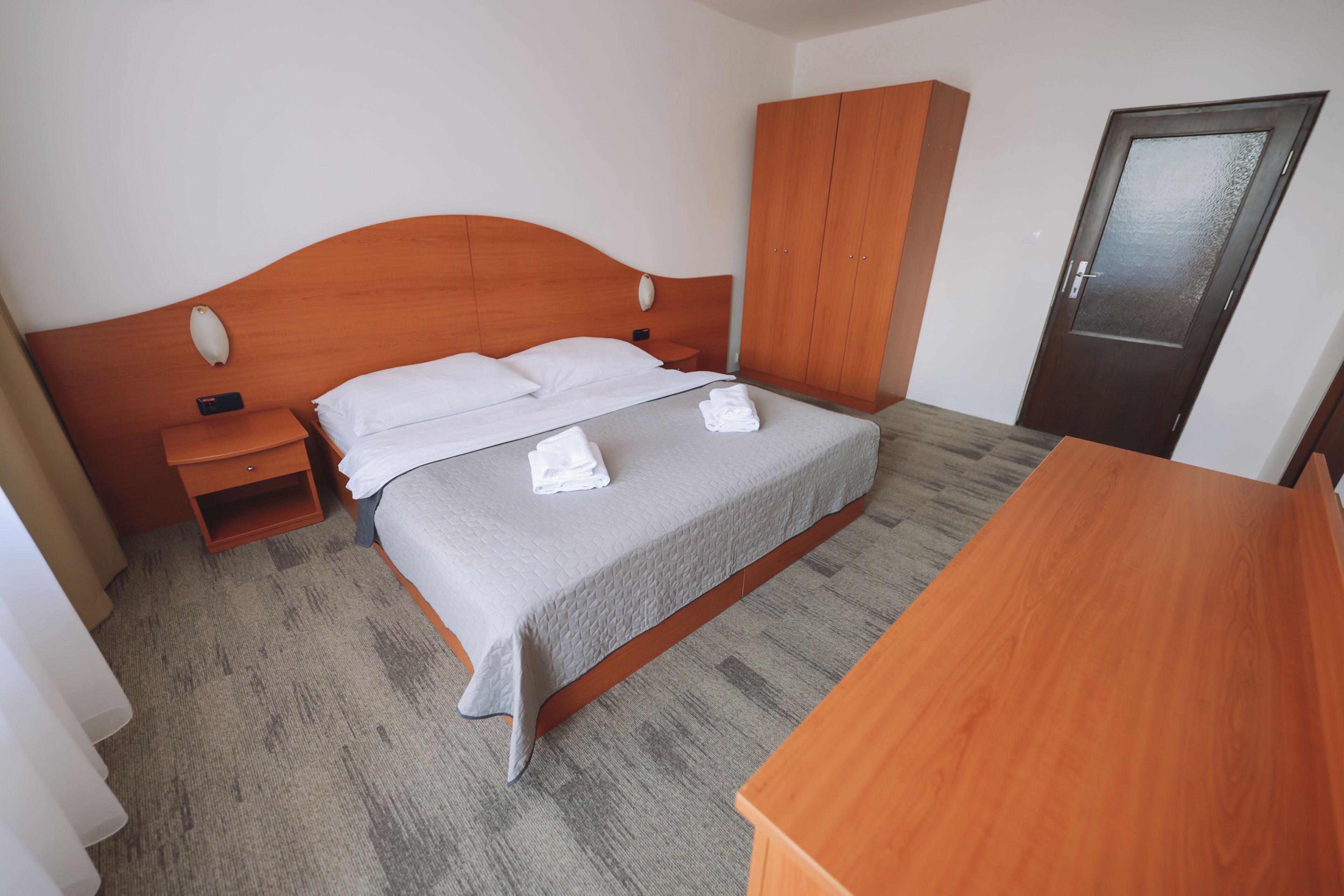 Hotellihuone, jossa on sänky, puinen yöpöytä, vaatekaappi ja ovi.