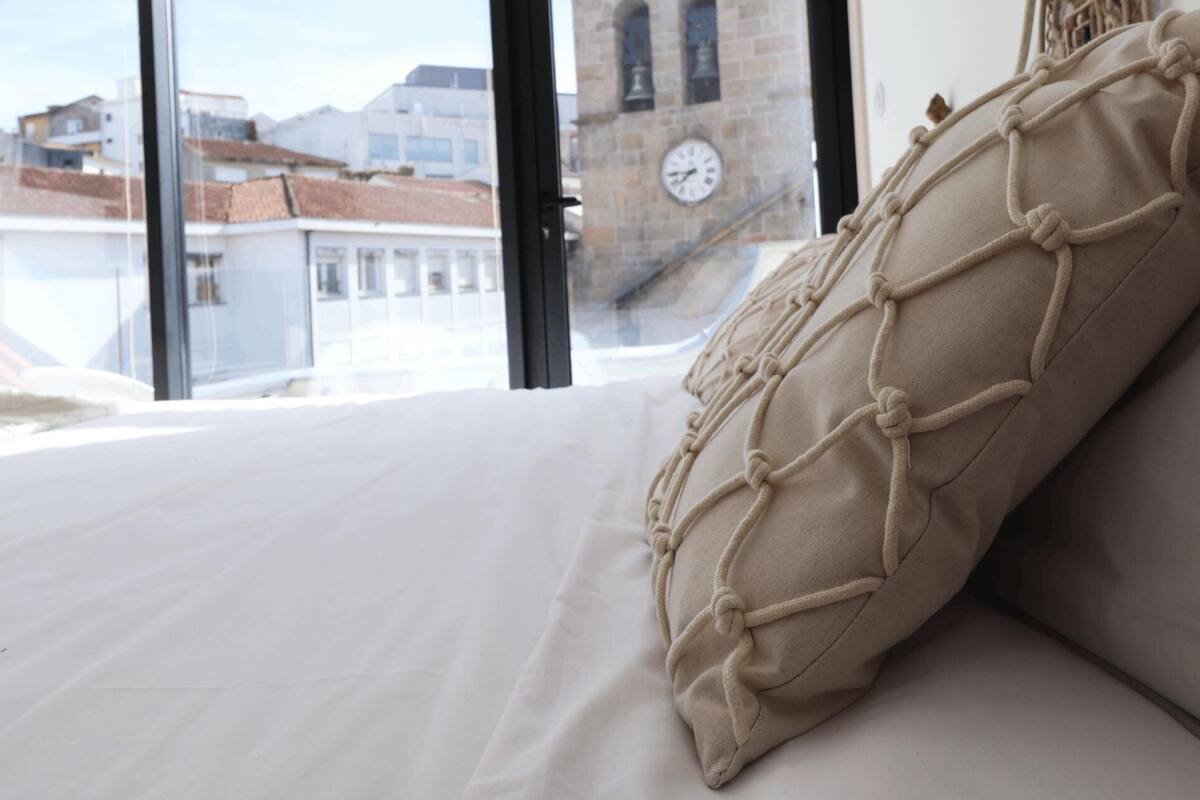 A bed with a beige macrame pillow and a view of buildings through a window.