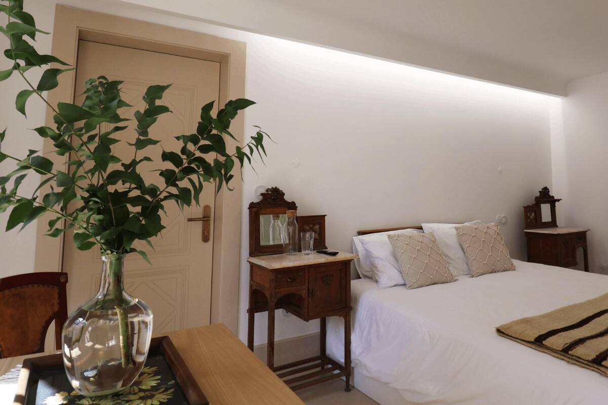 A bedroom with a bed, bedside tables, a nightstand, a wooden chair, a vase with a plant, and a window.