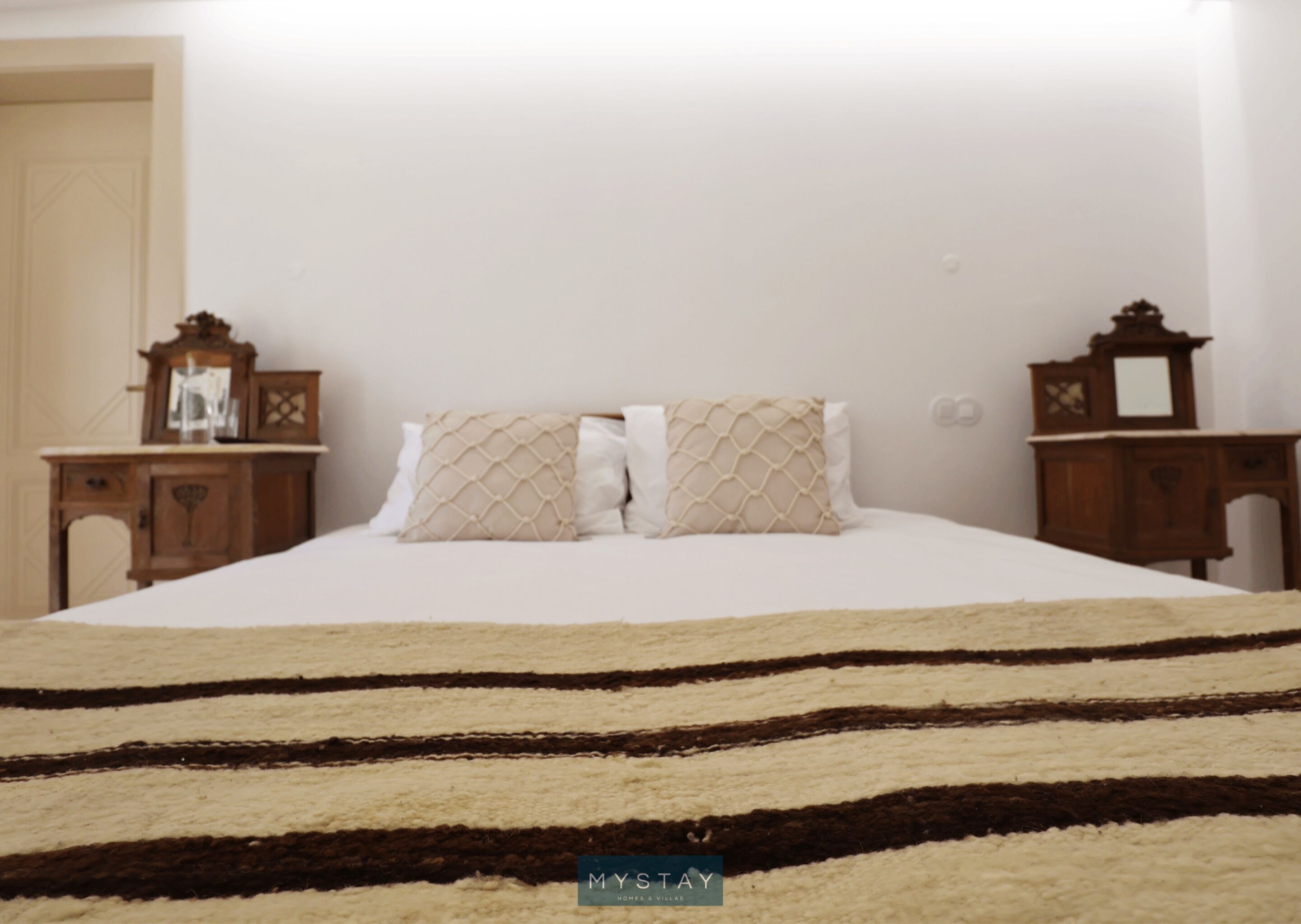 A bed with a striped rug, two bedside tables with clocks, and a mirror on the wall.