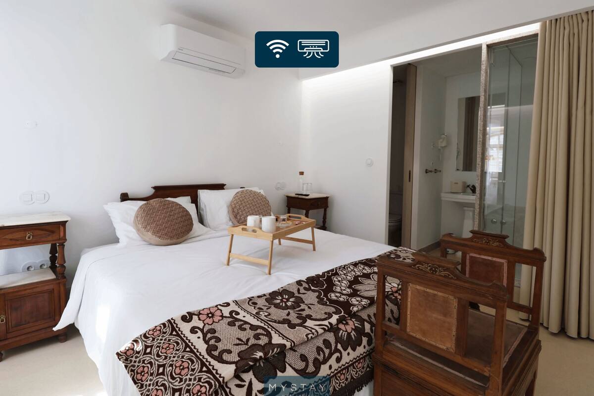 A hotel room with a bed, bedside tables, a wooden chair, a bedside table with a tray, and a sliding glass door leading to a bathroom.