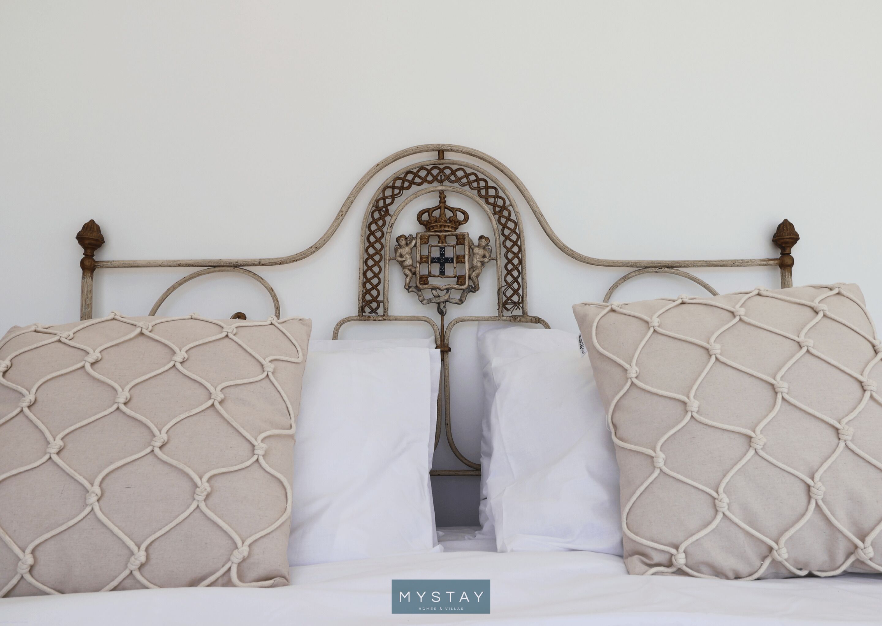 Una cama con cabecera de metal, dos almohadas color beige con diseño de mosquitero y una almohada blanca.