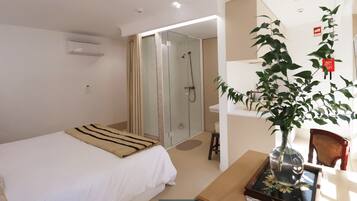 A hotel room with a bed, a bedside table, a small desk, a chair, a potted plant, a shower area, and a mirror.