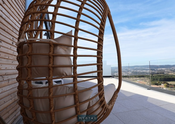 A wicker chair with cushions on a balcony overlooking a cityscape.