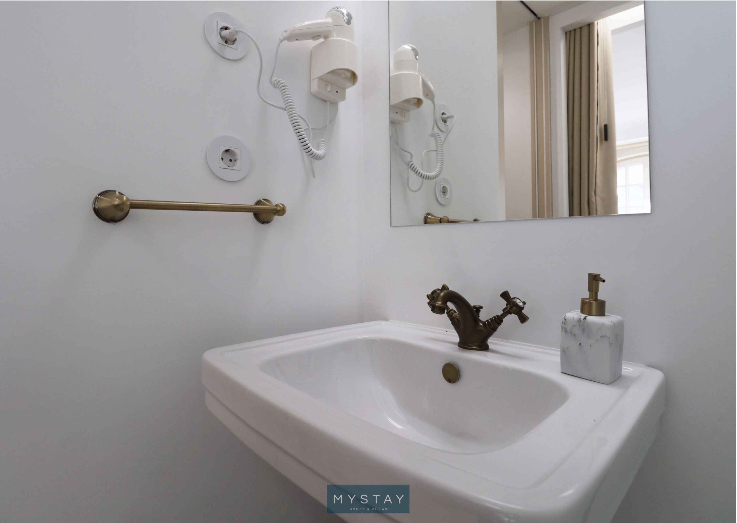 Un baño con un lavamanos blanco, un grifo de latón, un espejo y dos secadores de pelo montados en la pared.