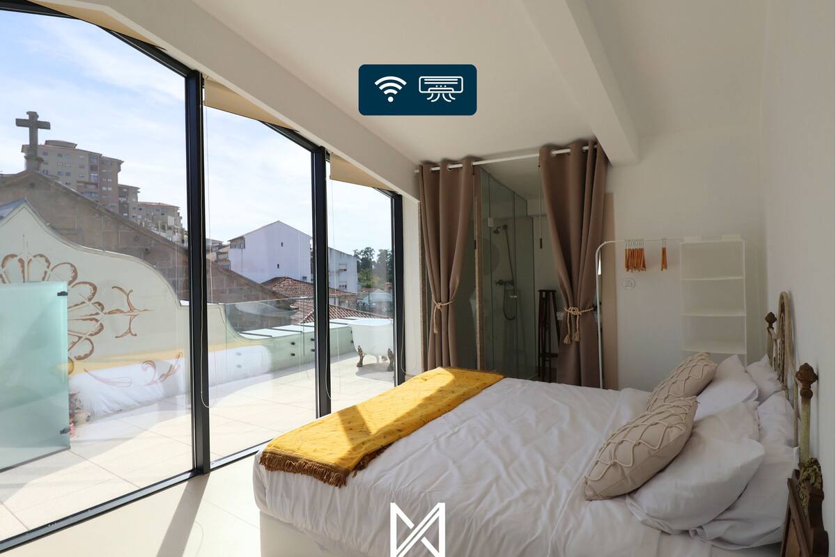 A bedroom with a large window, a bed with a yellow throw, and a view of buildings outside.