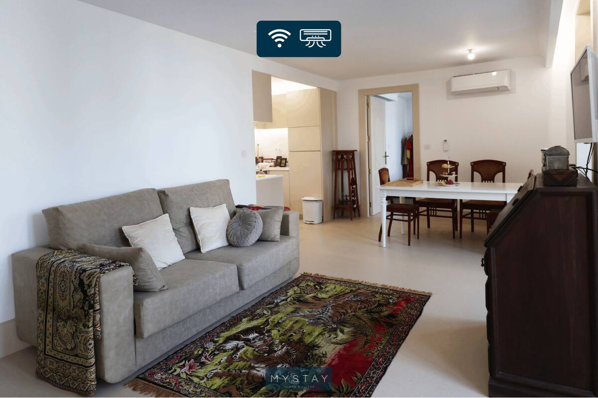 A living room with a sofa, a dining table, and chairs. There is a kitchen in the background.