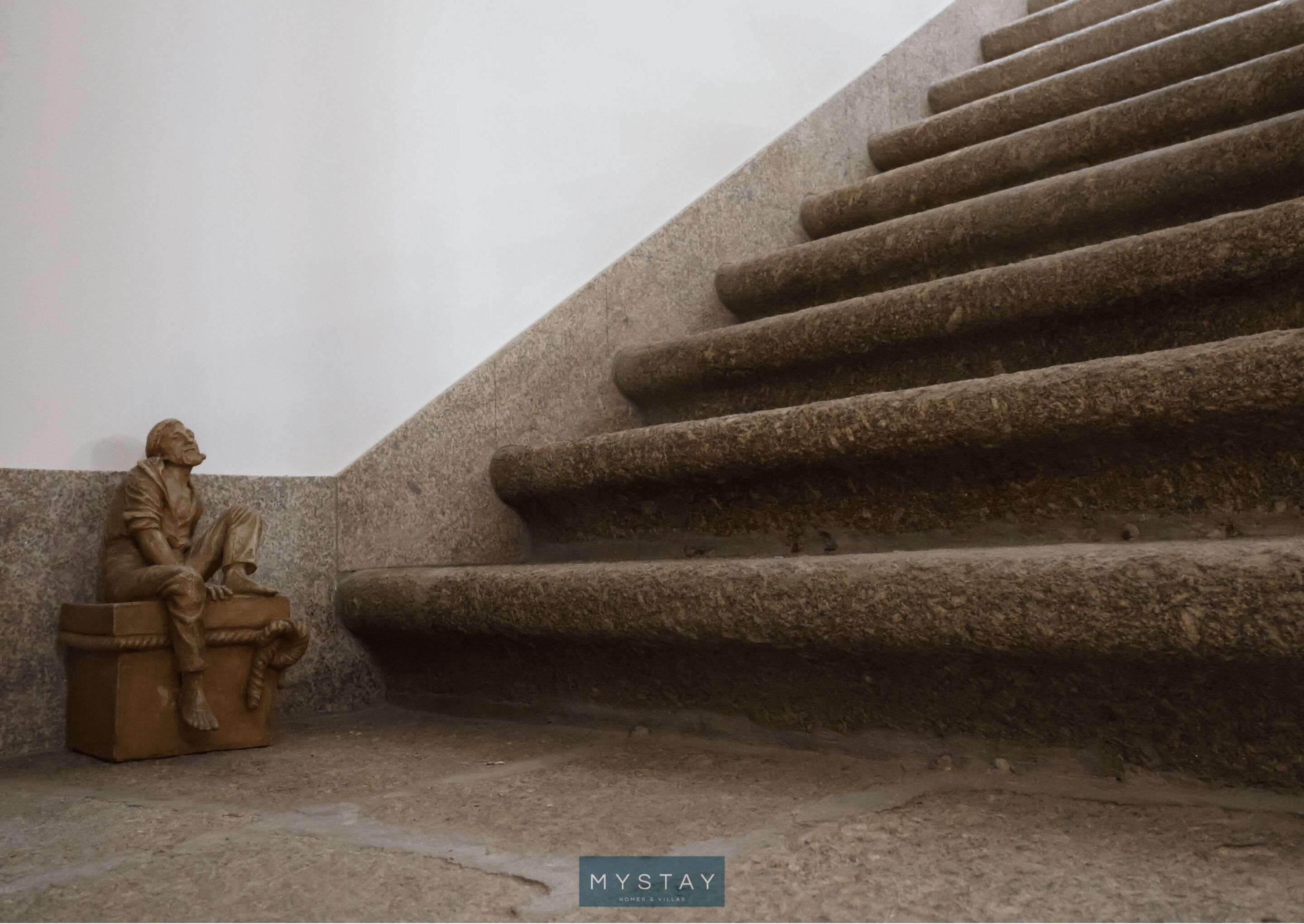 Una escultura de una figura sentada en un banco de piedra junto a una escalera.