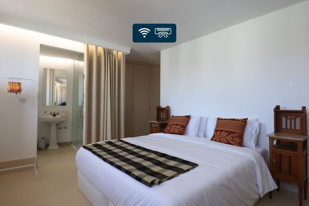 A hotel room with a large bed, a bedside table, a nightstand, a mirror, a wardrobe, and a bathroom with a sink and toilet.