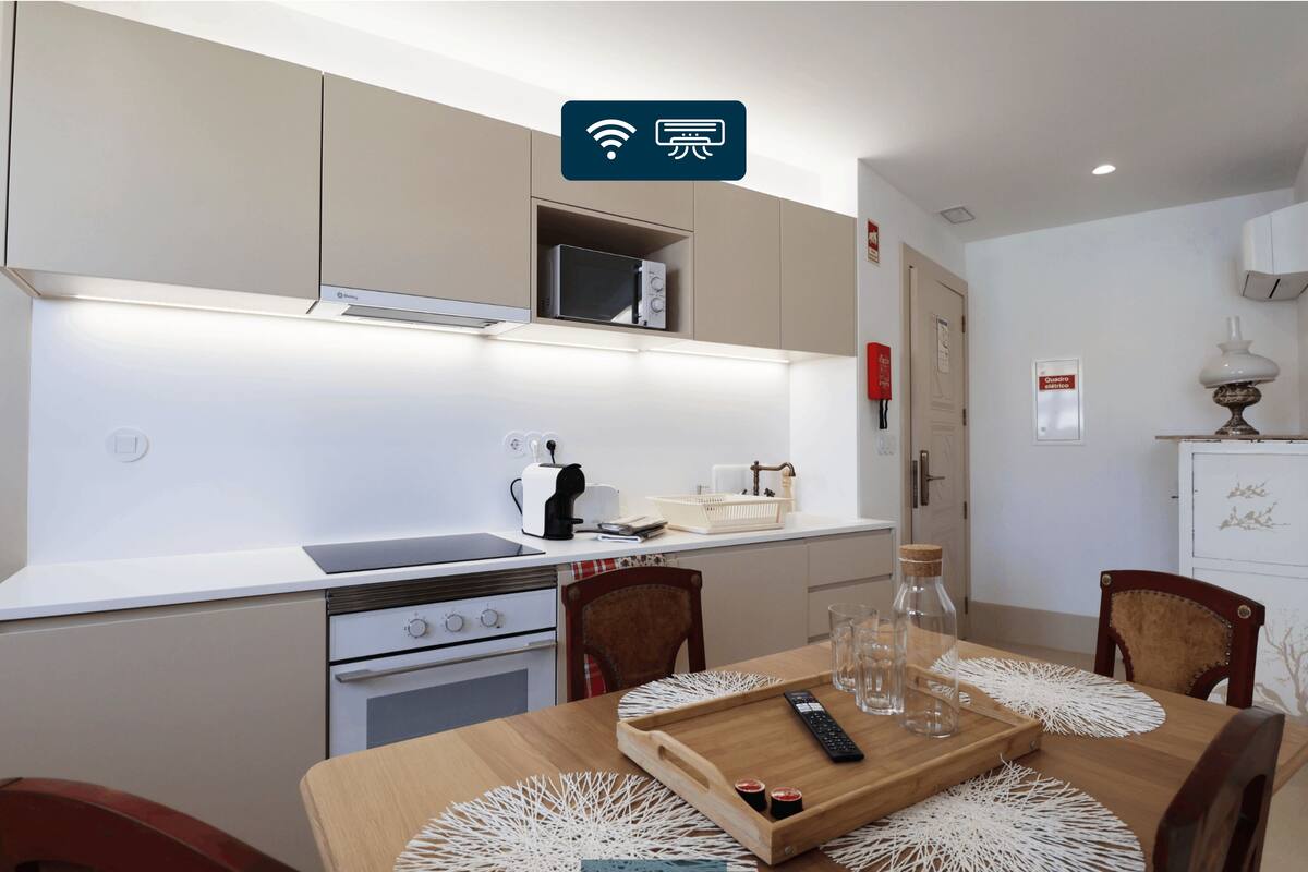 A modern kitchen with a microwave, toaster, and coffee maker on the countertop. There is a dining table with placemats and a tray with glasses and a remote control.