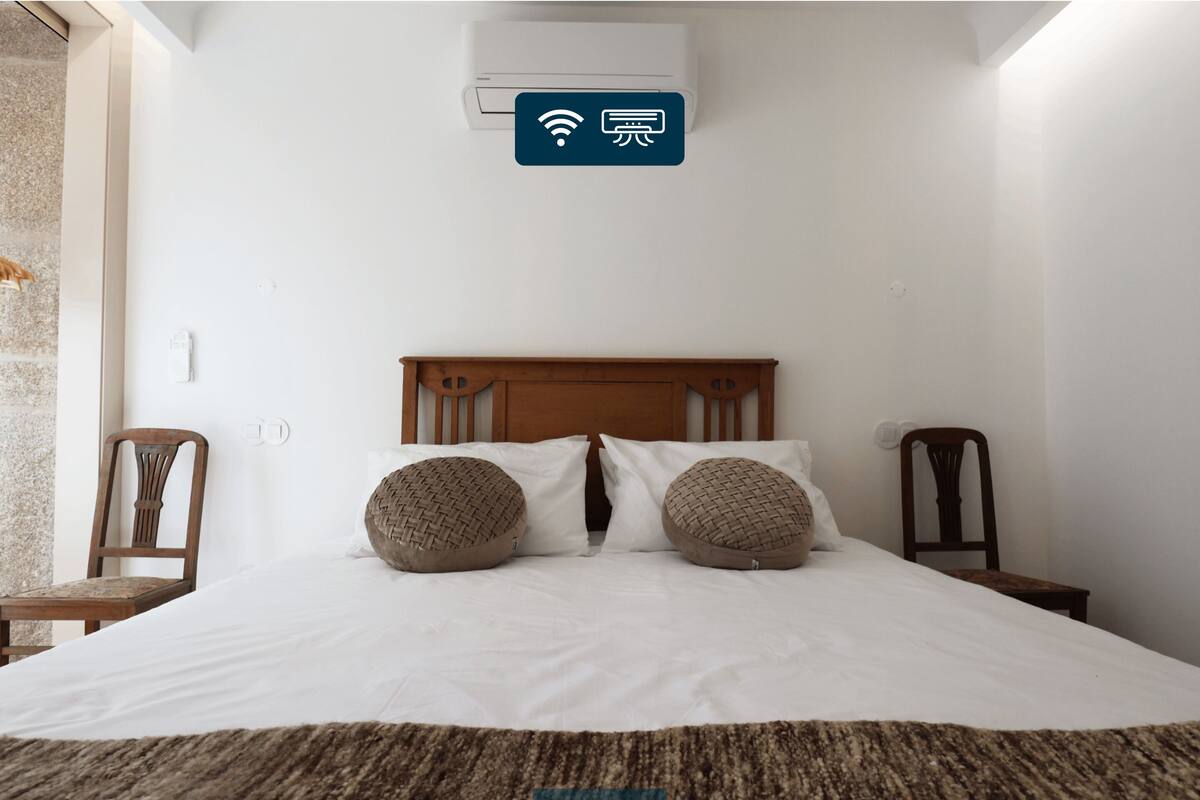 A bedroom with a bed, two pillows, a wooden headboard, a wall-mounted air conditioning unit, and a small table with a chair.