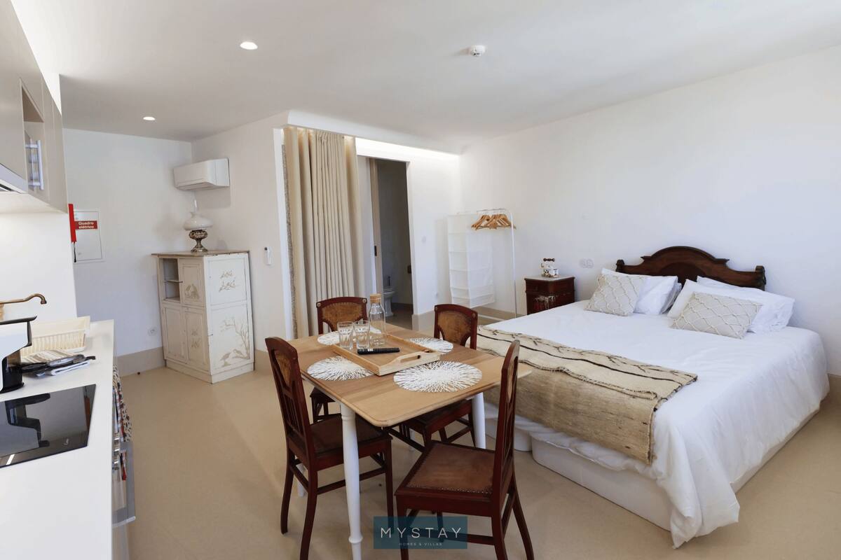 A hotel room with a bed, a dining table with chairs, a desk with a computer, a wardrobe, and a bathroom visible through a door.