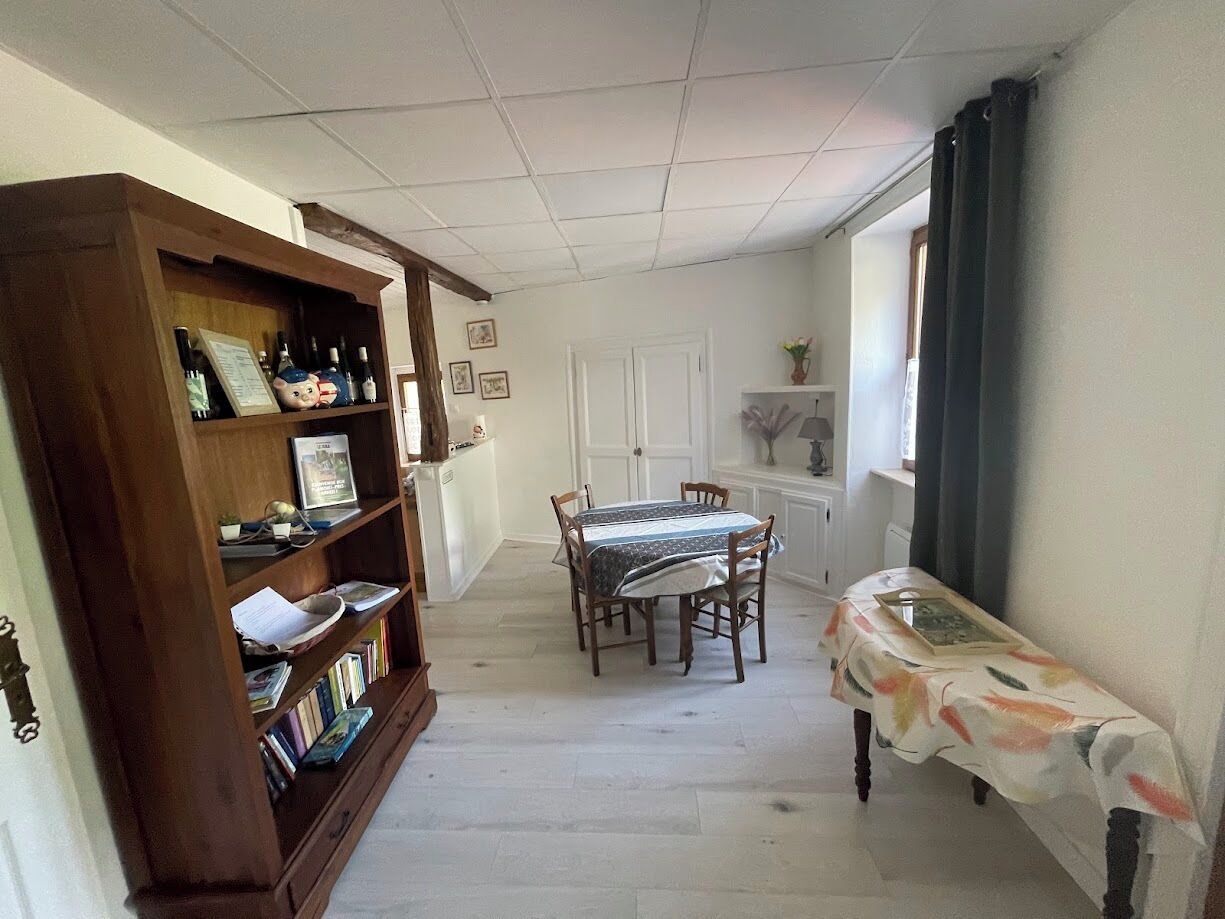 Een smalle gang met een houten boekenkast, een eettafel met stoelen en een kleine tafel met een bloemrijk tafelkleed.