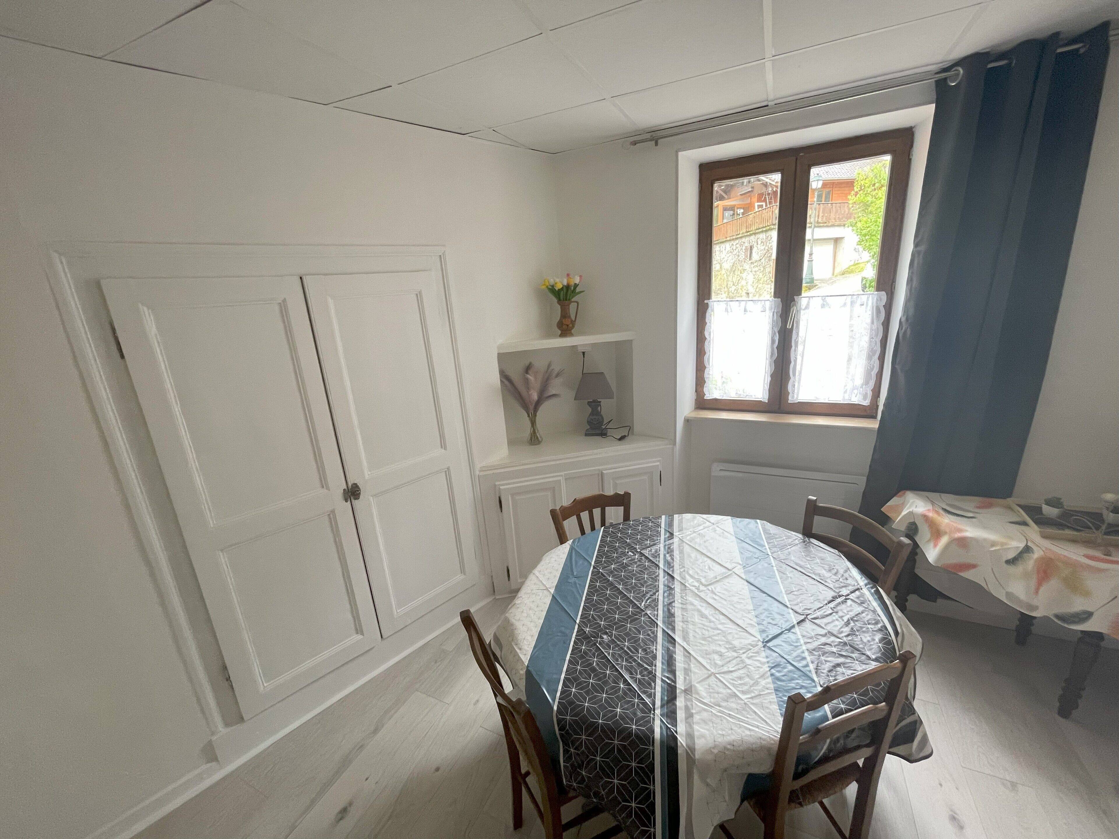 Een eetgedeelte met een ronde tafel, overdekt met een tafelkleed met dessin, omringd door houten stoelen. Er is een raam met uitzicht op groen, en een klein schap met decoratieve objecten.