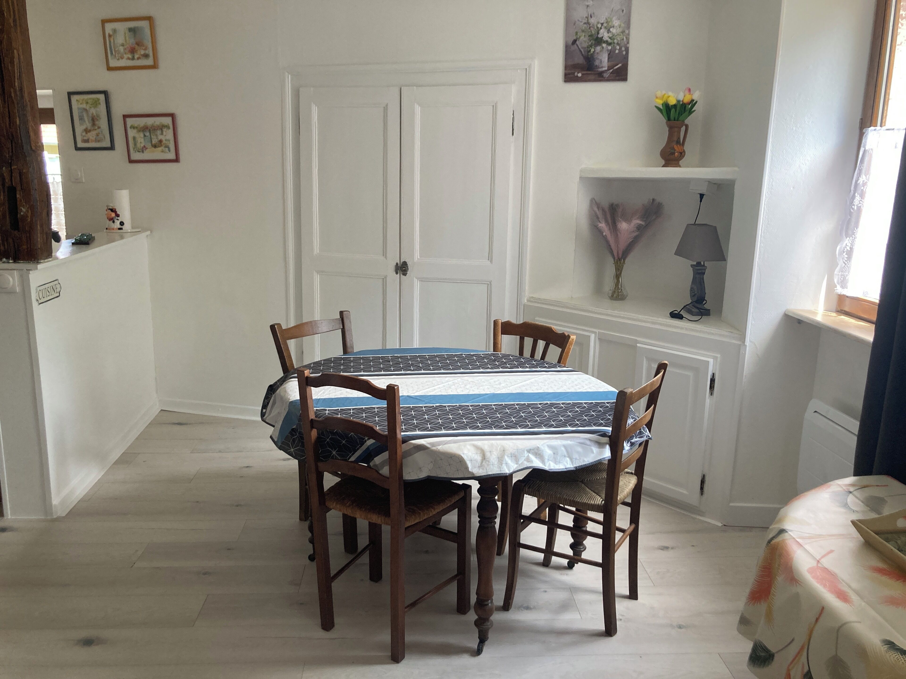 A dining area with a round table, four wooden chairs, a white door, and a small shelf with a lamp and flowers.