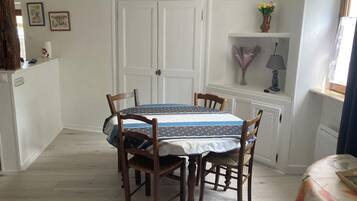 A dining area with a round table, four wooden chairs, a white door, and a small shelf with a lamp and flowers.