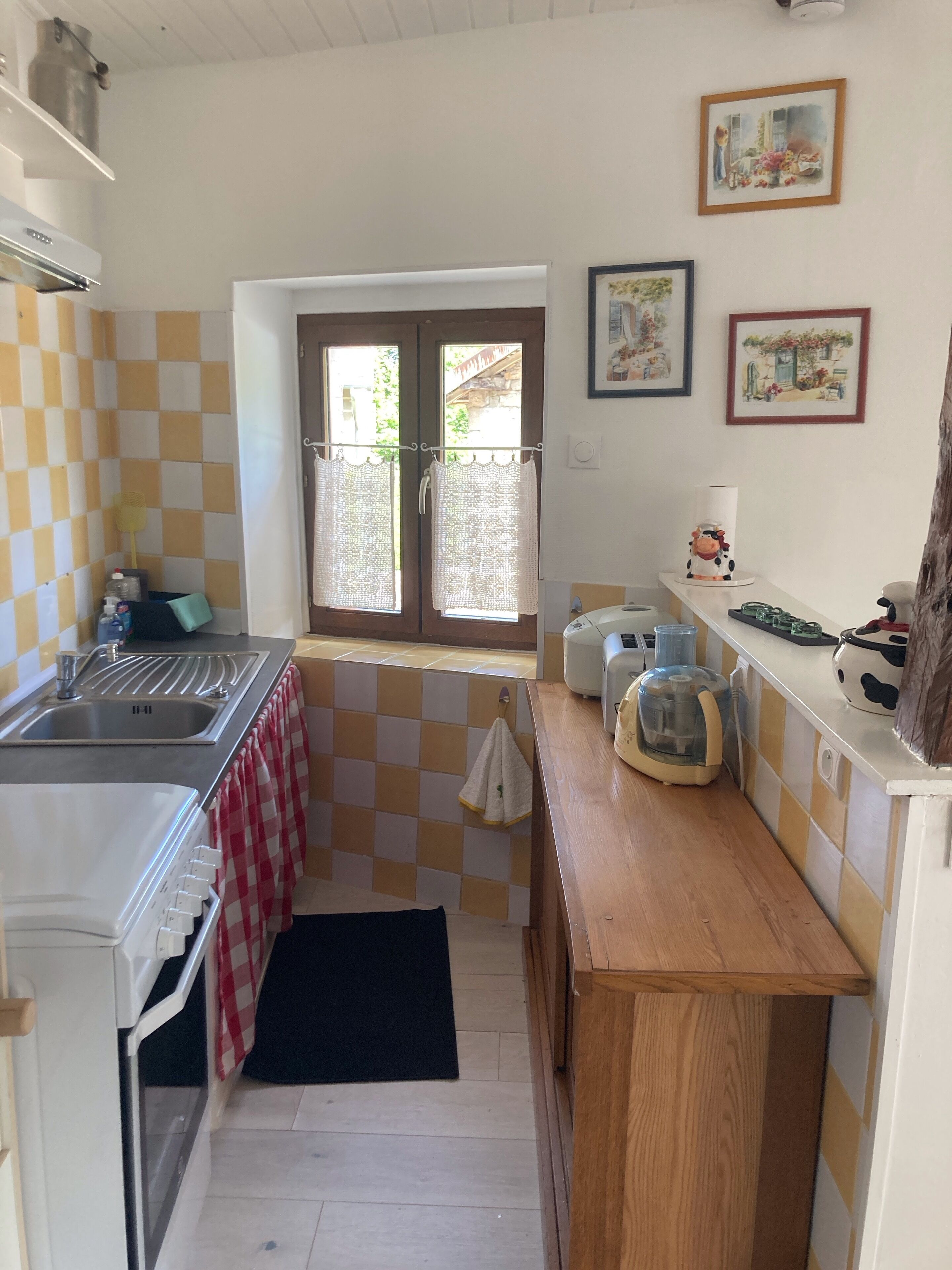 A compact kitchen with a sink, a stove, and a window with curtains. There are framed pictures on the wall and a small countertop with a coffee maker.