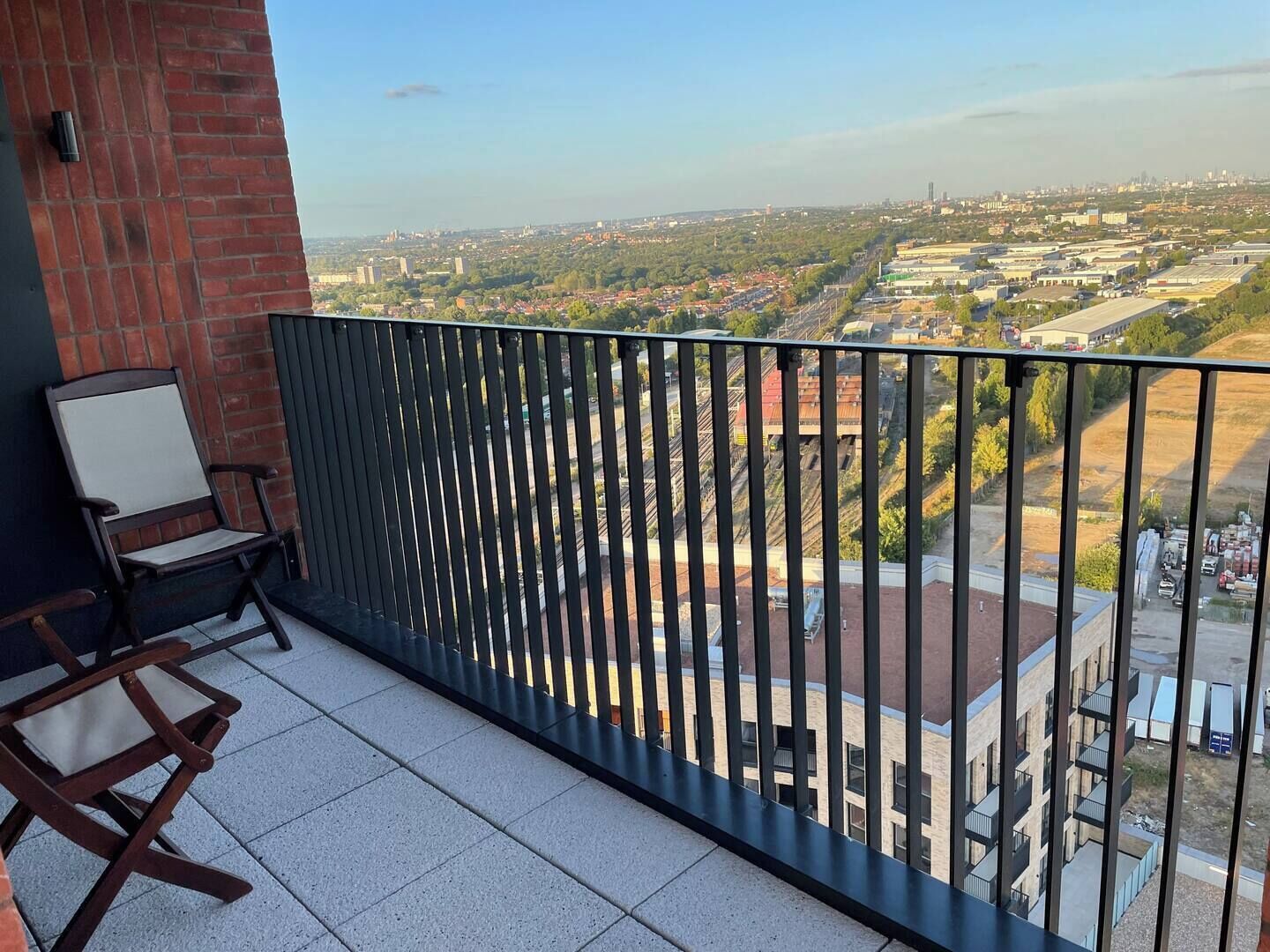 Un balcón con vistas a un paisaje urbano, un muro de ladrillo y dos sillas de madera.