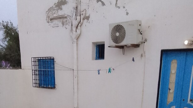 Ein Gebäude mit einer blauen Tür, einem kleinen Fenster mit blauem Rollo und einer an der Wand befestigten Klimaanlage.