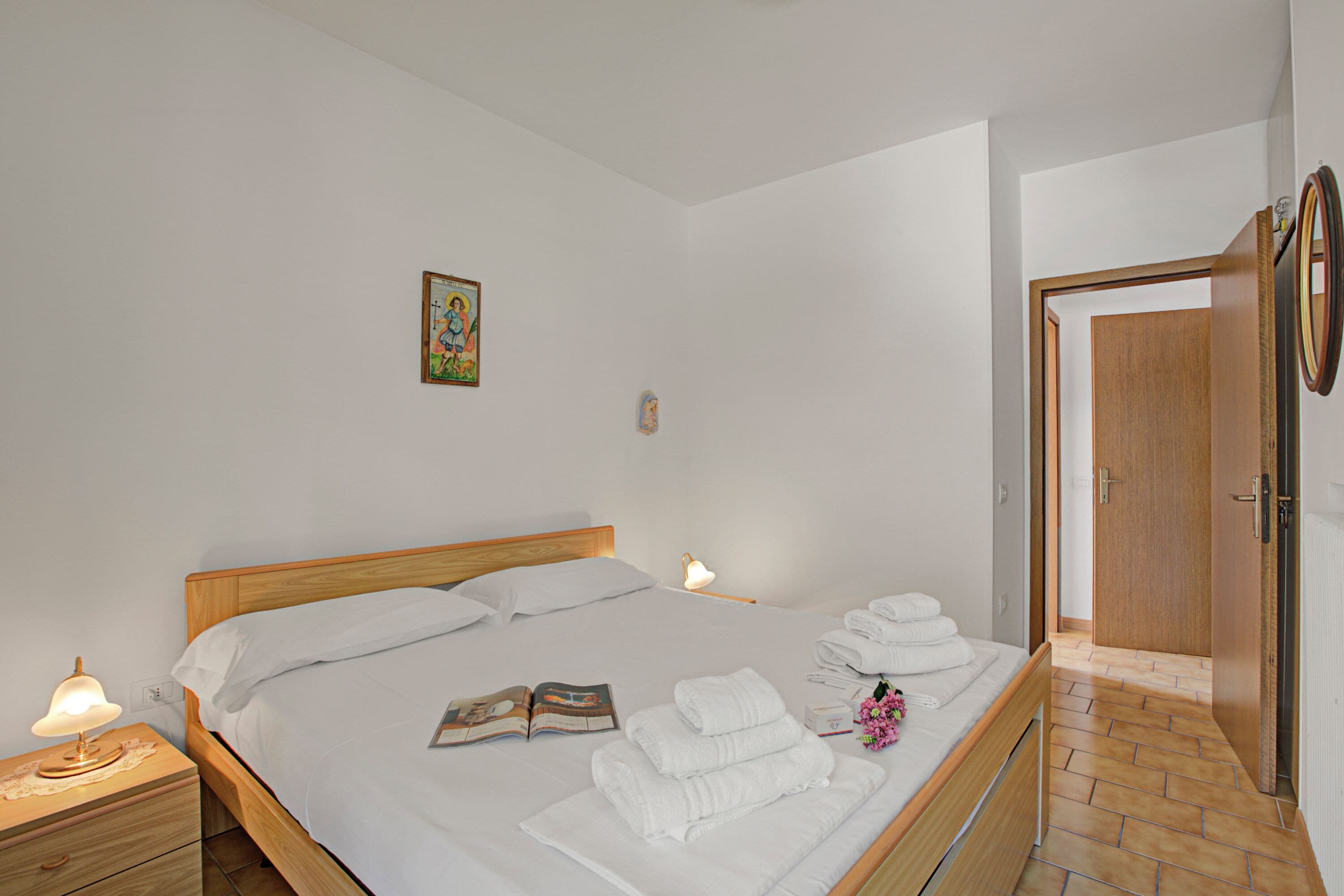 Una cama bien hecha con sábanas blancas, una lámpara de noche, un libro y toallas dobladas sobre un cabecero de madera.