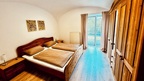 A bedroom with a wooden bed, a nightstand, a radiator, and a view of the outdoors through a sliding door. - Bauer Wirt`s Zwei-zimmer-wohnung (Kirchham)
