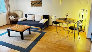 A cozy living room with a sofa, a dining table, and chairs. There's a wicker chair, a floor lamp, and a radiator. - Bauer Wirt`s Zwei-zimmer-wohnung (Kirchham)
