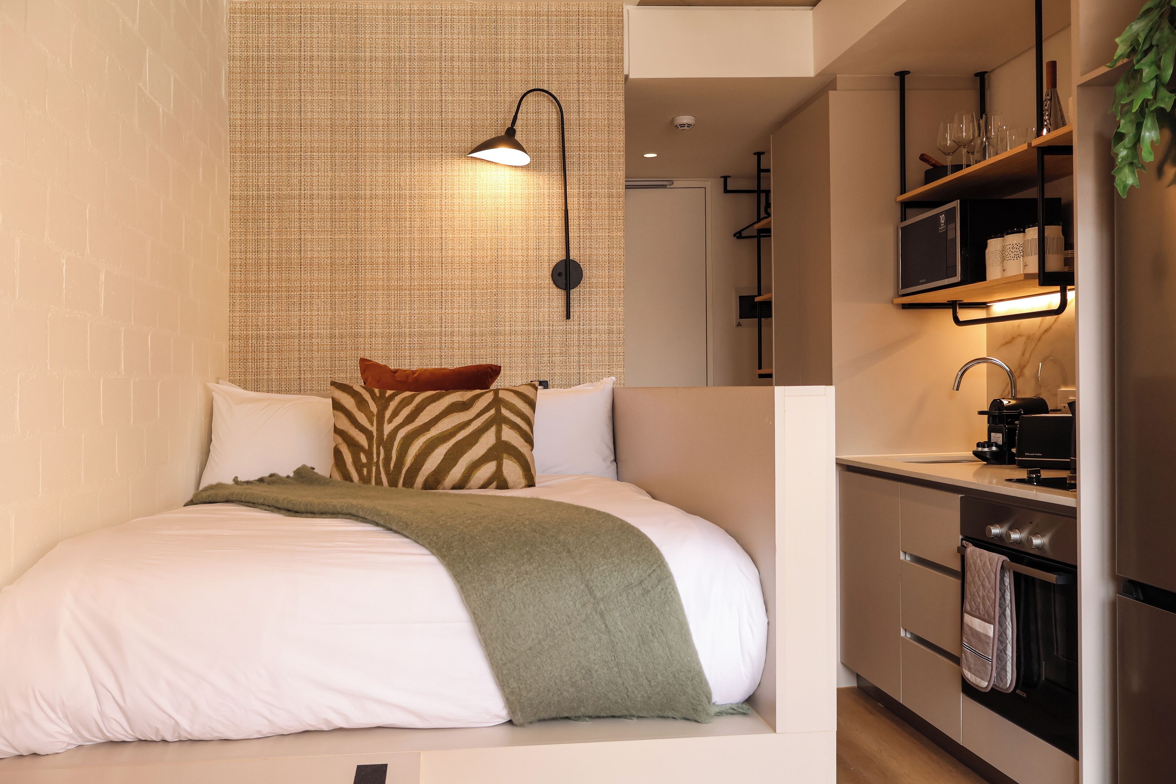 Een moderne slaapkamer met een groot bed, een groene plaid en een kussen met een zebraprint. Naast het bed bevindt zich een kitchenette met een koffieapparaat, een spoelbak en een fornuis.