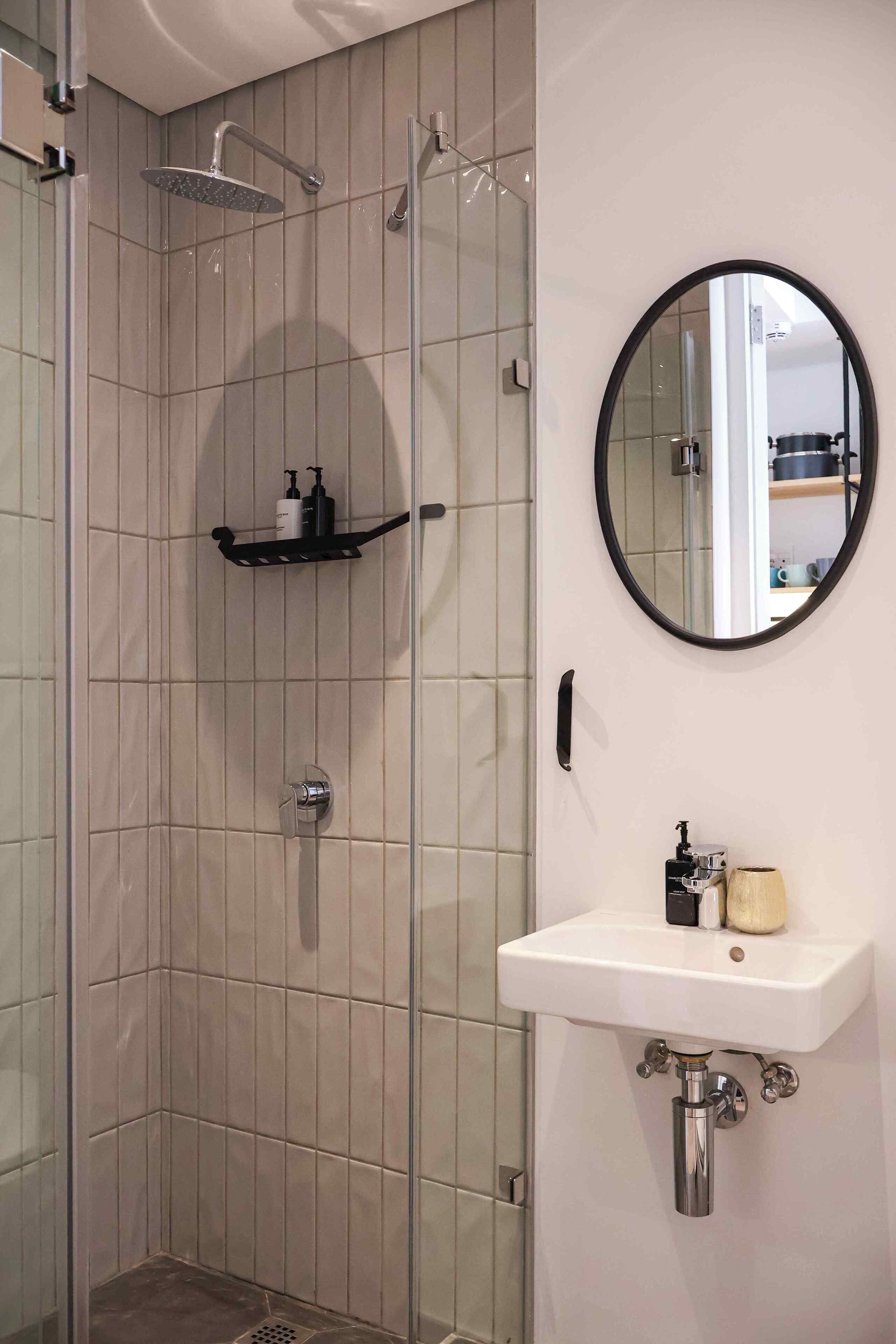 A modern bathroom with a glass-enclosed shower, a white sink with a round mirror, and a shelf with toiletries.