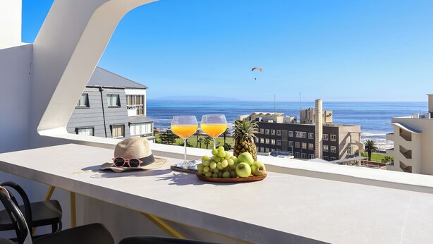 Een balkon met uitzicht op de oceaan, een tafel met een hoed, zonnebril, een schaal fruit en twee glazen jus d'orange.