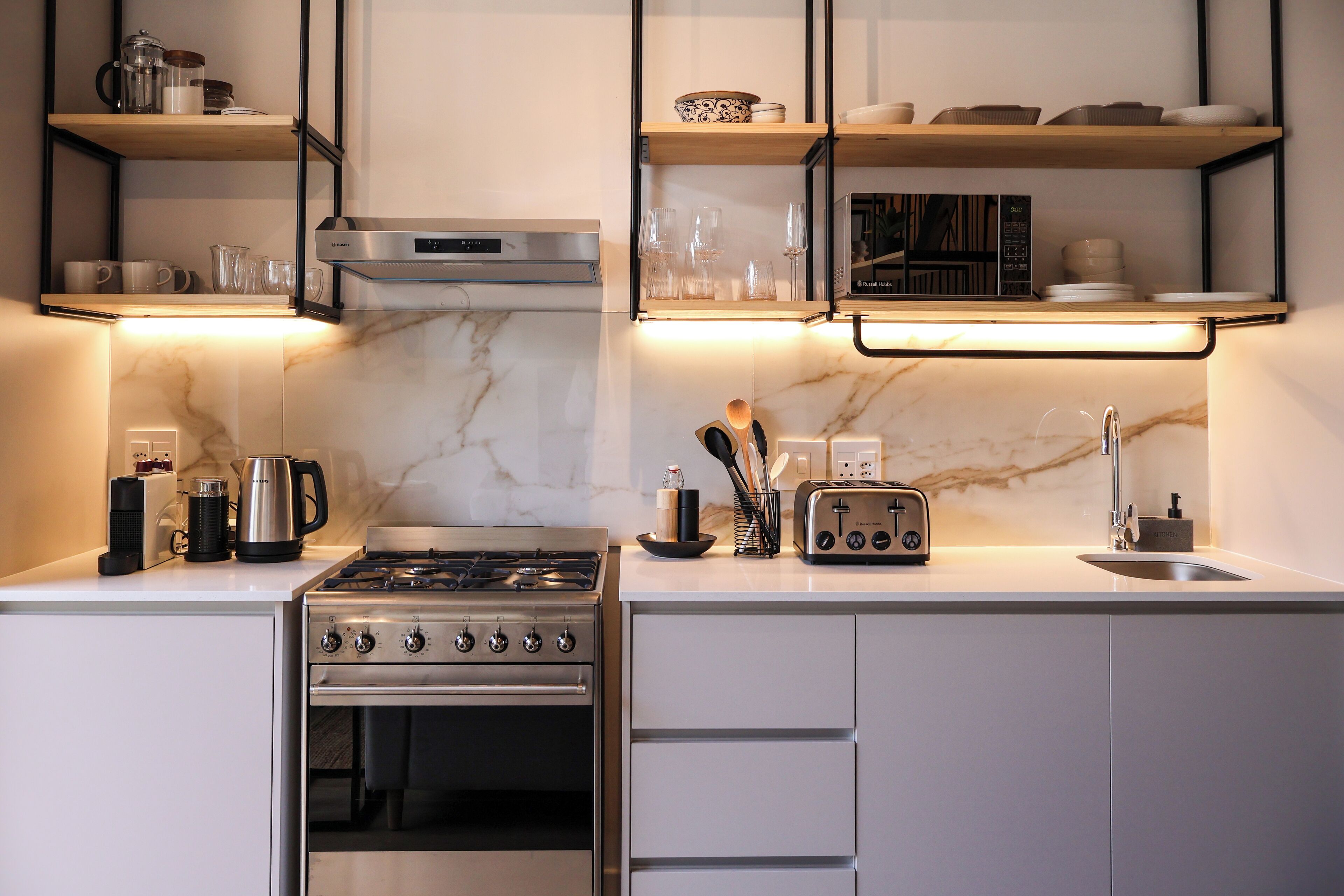 A modern kitchen with a gas stove, oven, and built-in toaster. There are wooden shelves above the counter holding dishes and glasses.