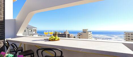 A balcony with a white railing, black chairs, and a table with fruit and drinks. A clear view of the ocean and buildings is visible.