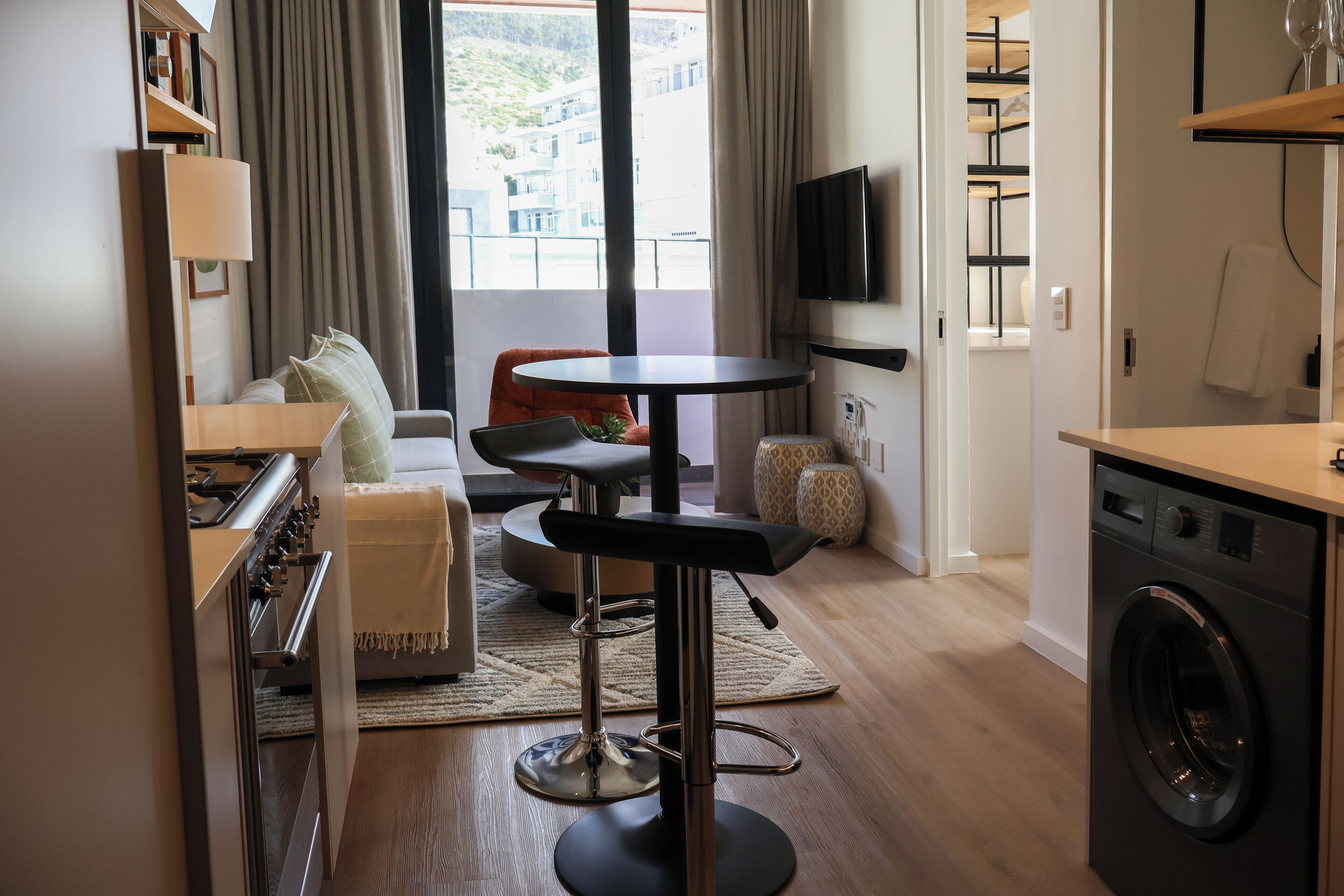 A modern kitchen with a washing machine, a dining area with a round table and chairs, and a living area with a sofa and a TV.